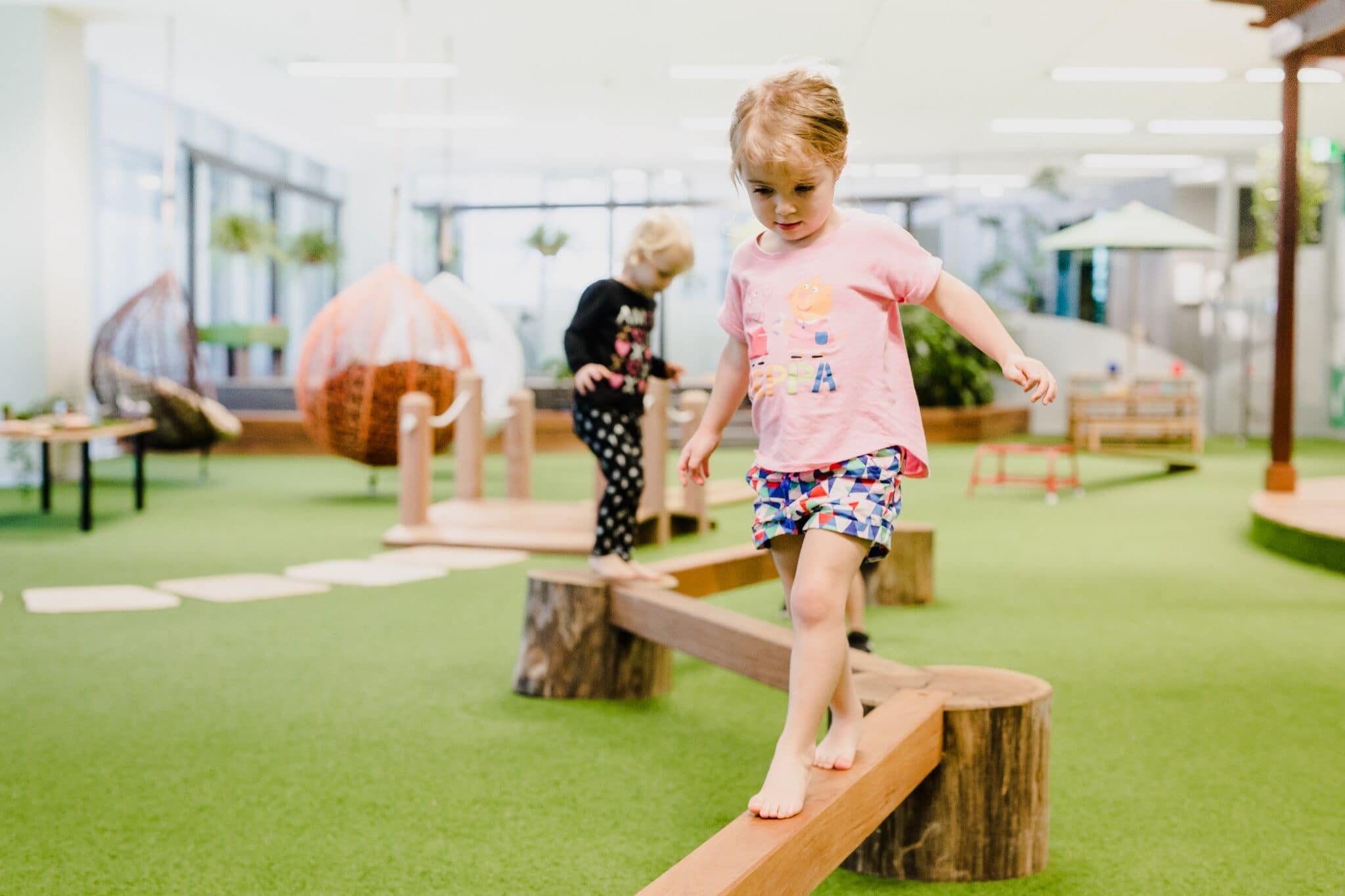Young child balancing on a wooden beam in an indoor play area with soft flooring, developing coordination and confidence.