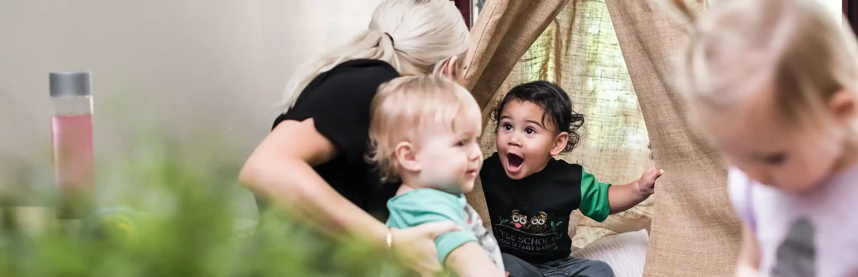Early childhood educator with two surprised and happy toddlers playing peek-a-boo near a canvas teepee in a cozy, sensory play area at Little Scholars.