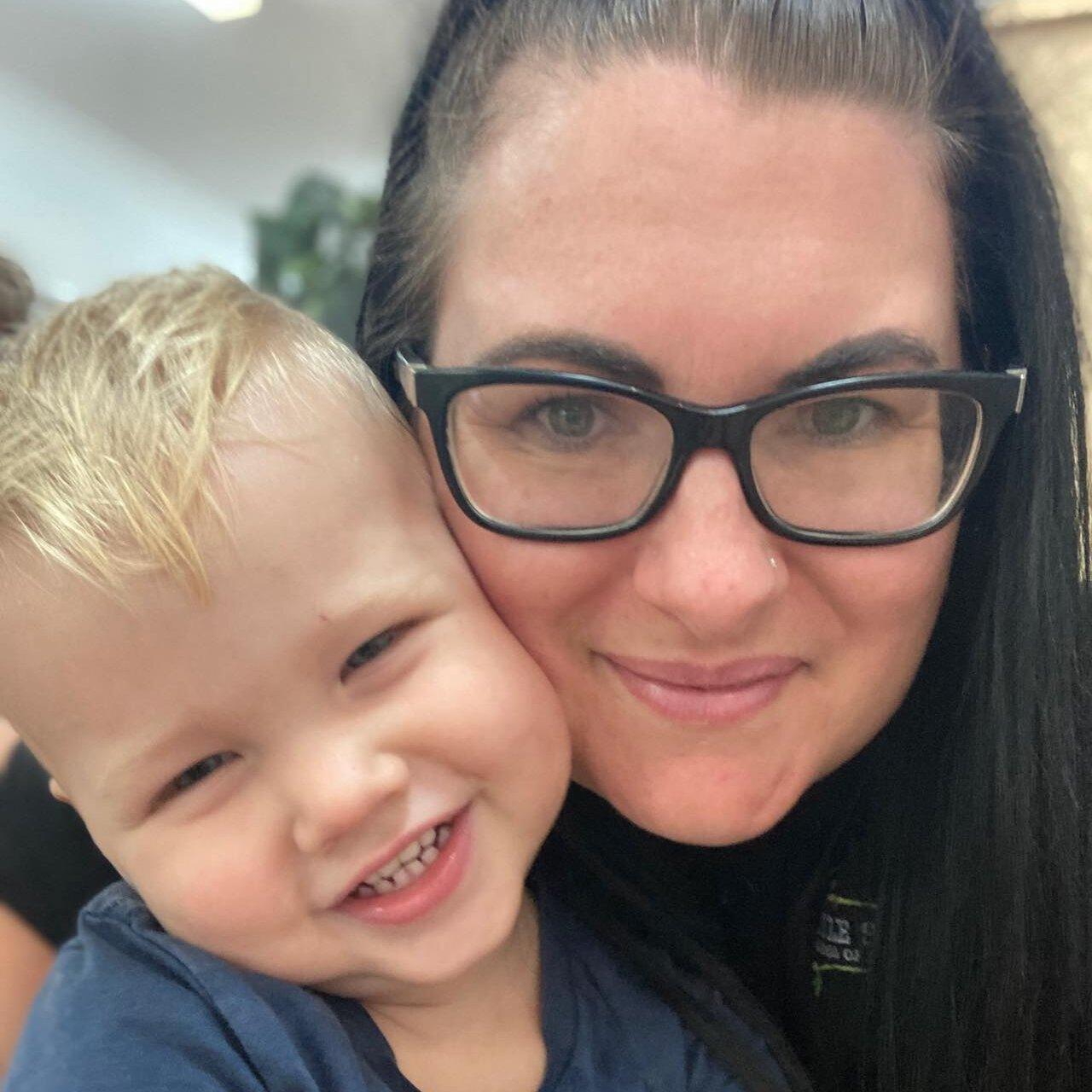 A loving close-up shows a smiling mother with dark hair and glasses cuddling a cheerful, blonde-haired toddler. The child leans into the mother's cheek, beaming widely, while the mother looks directly at the camera with a gentle expression. This heartwarming interaction visually complements content offering tips for separation anxiety in children.