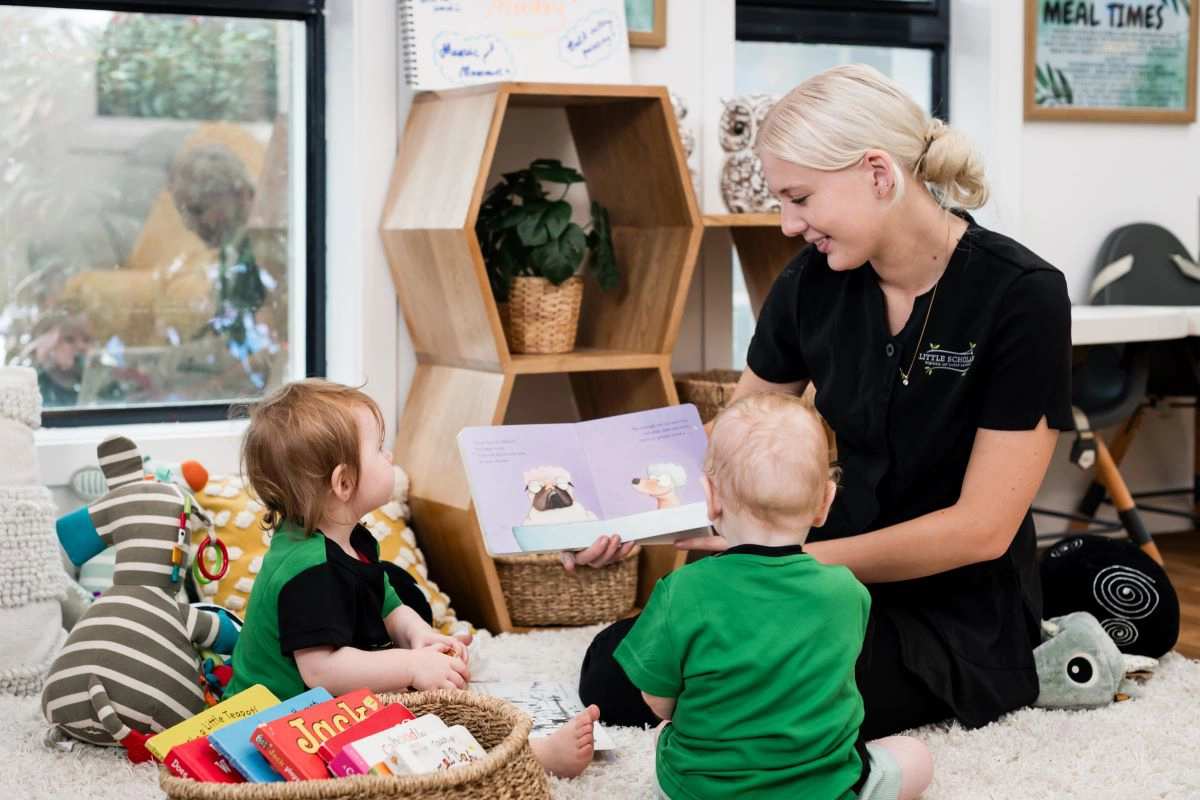 Two toddlers sit attentively on the floor listening to a Little Scholars educator reading a colorful picture book during story time, emphasizing early literacy and social-emotional development in the preschool environment.