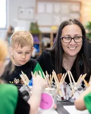 An educator smiles while supporting a child during an art activity, with coloured pencils and craft materials on the table in a classroom setting.